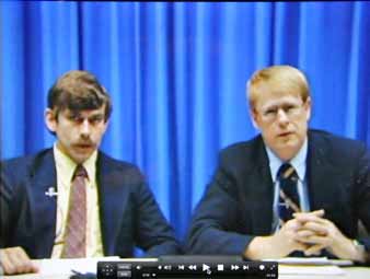 Two men in suits sitting in front of a blue curtain.