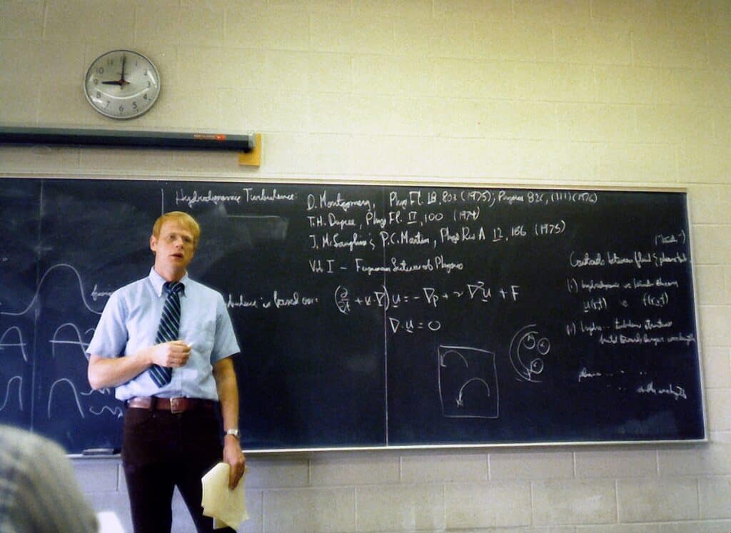 man in a suit in the 1970s standing in front of a chalkboard with equations drawn on it