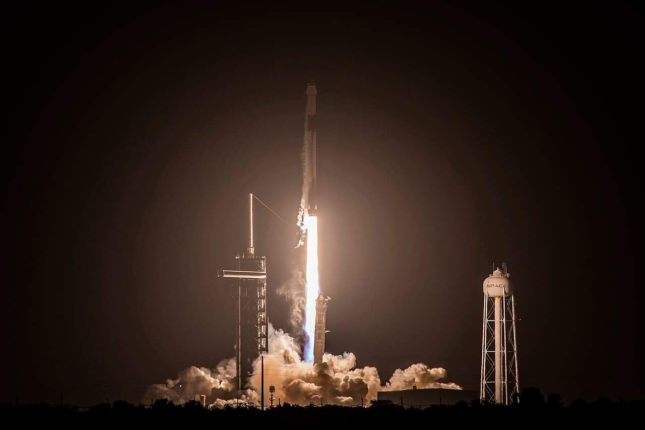 A SpaceX Falcon 9 rocket launches into the night sky from Kennedy Space Center, leaving a bright trail of flames and smoke against the dark backdrop. A water tower with the SpaceX logo stands illuminated to the right of the launch pad.