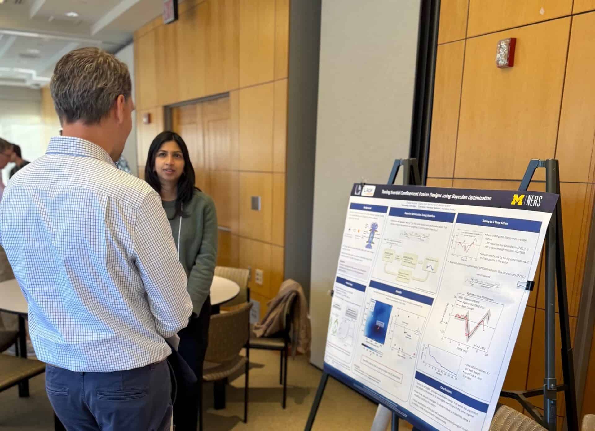 A woman presents a research poster to a man at an indoor academic event. The poster is titled “Tuning Inertial Confinement Fusion Designs using Bayesian Optimization” and features charts, diagrams, and text. The University of Michigan and NERS (Nuclear Engineering and Radiological Sciences) logos are visible on the poster. The woman is speaking while the man listens attentively, standing with his back to the camera. The setting is a modern conference room with wood-paneled walls and round tables.