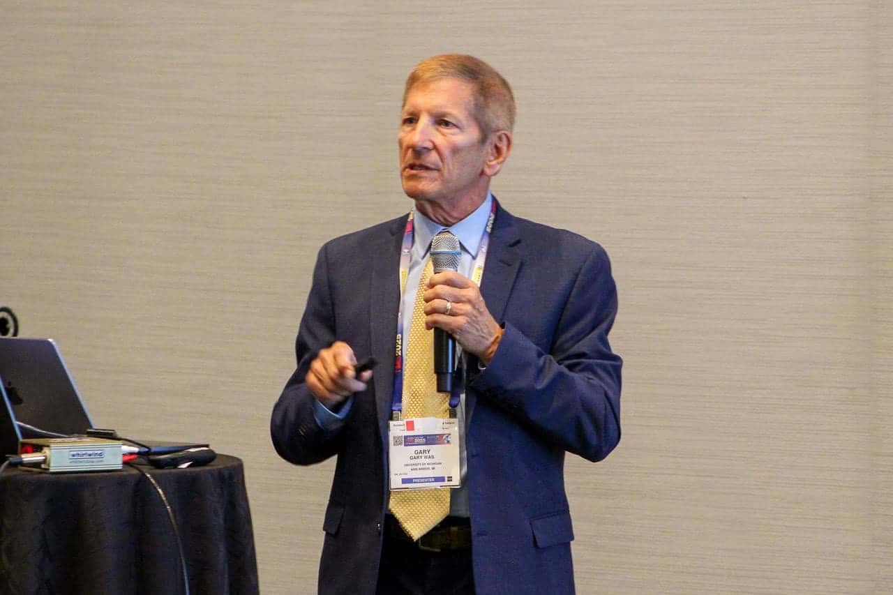 Gary Was stands at the front of a conference room giving a presentation. He is wearing a navy suit with a yellow tie and holding a microphone in one hand and a presentation remote in the other. A name badge with “PRESENTER” is visible around his neck.